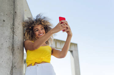 Lächelnde junge Frau, die ein Selfie mit ihrem Smartphone an der Wand macht - PGF00625