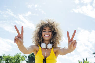 Glückliche junge Afro-Frau, die an einem sonnigen Tag ein Friedenszeichen macht - PGF00649