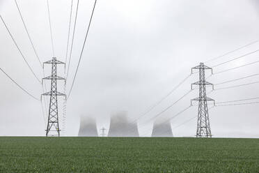 UK, England, Rugeley, Electricity pylons standing in field during foggy weather with cooling towers in background - WPEF04930