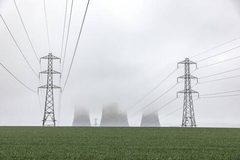 UK, England, Rugeley, Electricity pylons standing in field during foggy weather with cooling towers in background - WPEF04930