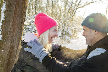 Cheerful senior couple wearing knit hats looking at each other in forest - FVDF00286