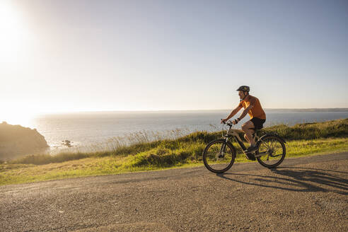 Mature sportsman cycling bicycle on road near sea at sunset - UUF23732