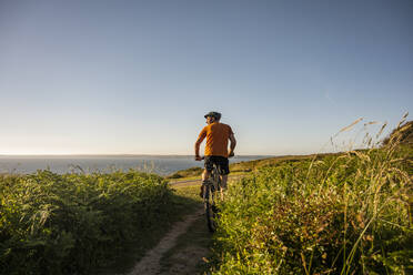 Älterer männlicher Sportler fährt elektrisches Mountainbike inmitten von Pflanzen bei Sonnenuntergang - UUF23742