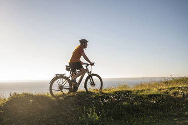 Male sportsperson riding electric mountain bike on green grass at sunset - UUF23746