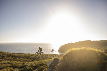 Männlicher Sportler fährt mit einem elektrischen Mountainbike auf einer Klippe am Meer - UUF23755