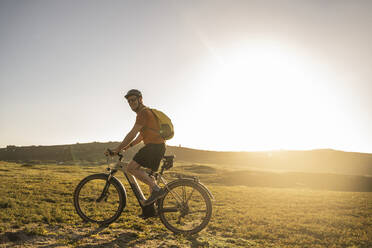 Mature sportsman with backpack riding electric bicycle at sunset - UUF23763