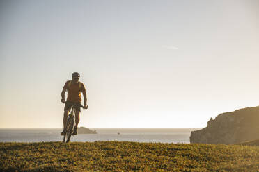 Männlicher Sportler fährt Mountainbike auf einer Wiese bei Sonnenuntergang - UUF23764