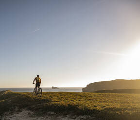 Männlicher Sportler auf einem elektrischen Mountainbike auf einer Wiese bei Sonnenuntergang - UUF23765