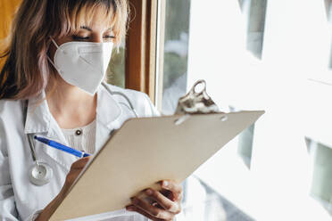 Ärztin mit Gesichtsschutzmaske schreibt in einer Klinik auf einem Klemmbrett - EGHF00054