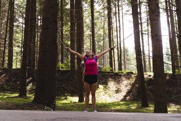Carefree female hiker with arms outstretched holding hiking poles while standing in forest - FMOF01441