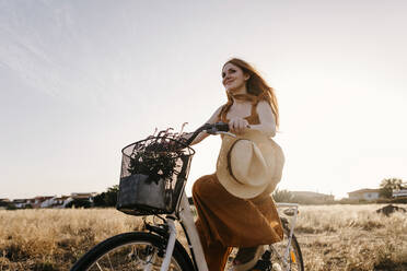 Redhead woman riding bicycle in field during sunset - TCEF01968