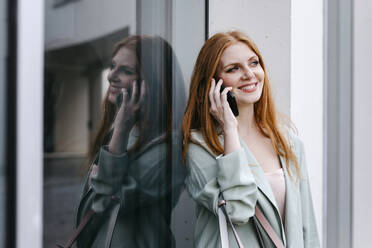 Rothaarige weibliche Fachkraft, die wegschaut, während sie über ihr Smartphone spricht und sich an ein Fenster lehnt - TCEF01986