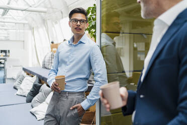 Männlicher Berufstätiger, der einen Einwegbecher hält, während er sich mit einem Kollegen im Büro unterhält - DIGF15905