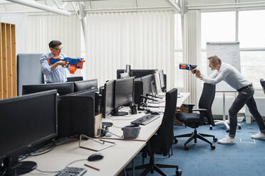 Männliche Kollegen spielen mit einer Spielzeugpistole, während sie am Schreibtisch stehen - DIGF15954