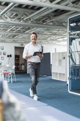 Smiling businessman holding digital tablet while walking in office - DIGF15960