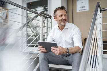 Lächelnder Geschäftsmann, der ein digitales Tablet hält, während er an einem Geländer im Büro sitzt - DIGF15974