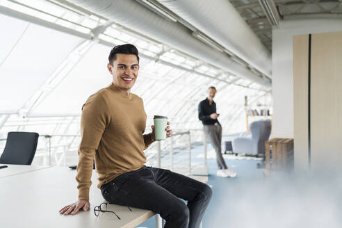 Lächelnder Geschäftsmann, der einen Einwegbecher hält, während er auf dem Schreibtisch sitzt und ein Kollege im Hintergrund im Büro steht - DIGF16004