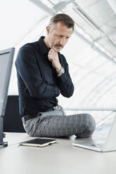 Male professional with hand on chin sitting by laptop in office - DIGF16016