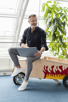 Lächelnder Geschäftsmann sitzt mit Laptop auf Auto-Spielzeug im Büro - DIGF16020