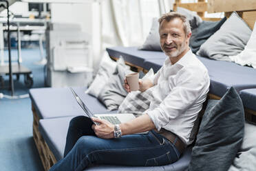 Smiling male professional with disposable cup and laptop sitting on sofa - DIGF16027