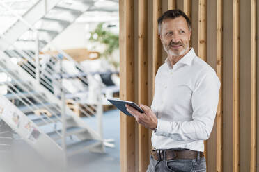 Nachdenklicher Geschäftsmann, der ein digitales Tablet hält, während er an der Wand im Büro steht - DIGF16038