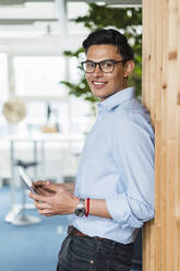 Businessman holding digital tablet while leaning on wall in office - DIGF16043