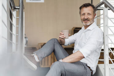 Lächelnder Geschäftsmann hält Kaffeetasse, während er auf einer Treppe sitzt - DIGF16054