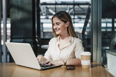 Smiling young businesswoman using laptop sitting at table in cafe - EBBF04091