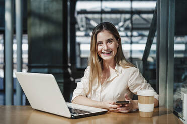 Happy female professional sitting with laptop and disposable coffee cup at table in cafe - EBBF04092