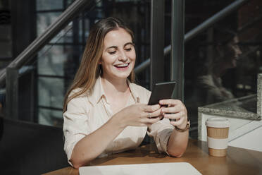 Glückliche junge Freiberuflerin, die in einem Café eine Textnachricht über ihr Smartphone verschickt - EBBF04093