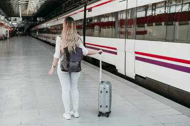 Junge weibliche Passagierin mit Rucksack steht mit Gepäck auf Rädern am Bahnsteig - EBBF04126