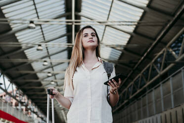 Schöne junge Frau mit Smartphone und Gepäck wartet am Bahnhof - EBBF04127