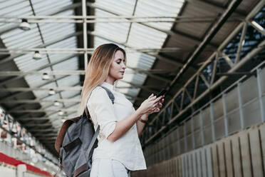 Young woman wearing backpack using smart phone at railroad station - EBBF04128