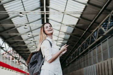 Cheerful young female passenger wearing backpack looking away at railroad station - EBBF04129