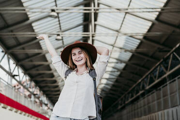 Glücklicher junger weiblicher Fahrgast mit Hut steht mit erhobenen Händen am Bahnhof - EBBF04131