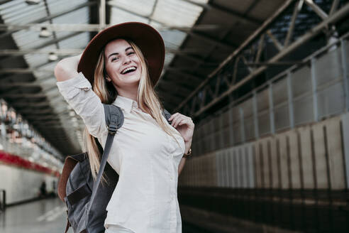 Fröhliche blonde Frau mit Rucksack am Bahnhof - EBBF04137