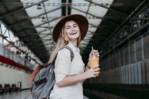 Glückliche junge Frau mit Hut und Einweg-Saftbecher auf dem Bahnhof - EBBF04140