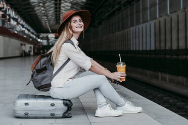 Lächelnder junger weiblicher Fahrgast mit Saft in der Hand, der auf einem Gepäckstück am Bahnsteig sitzt - EBBF04141