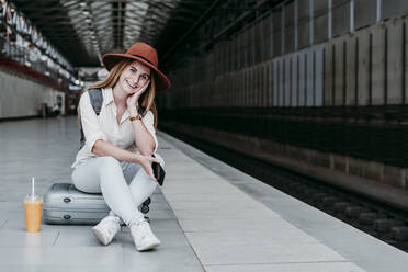 Glückliche junge Frau mit Hut auf einem Koffer sitzend auf dem Bahnsteig - EBBF04142