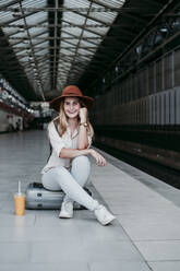 Glückliche Frau mit Hut, die auf einem Gepäckstück auf dem Bahnsteig sitzt - EBBF04143