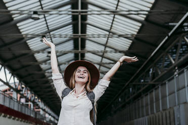 Fröhliche junge Frau, die mit erhobenen Händen am Bahnhof ihren Urlaub genießt - EBBF04144