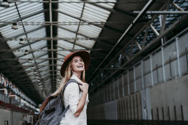 Cheerful young female passenger wearing backpack looking away at railroad station - EBBF04145