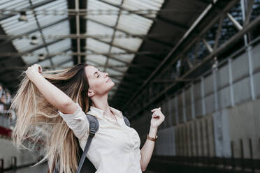 Beautiful young woman with eyes closed holding long blond hair at railroad station - EBBF04146