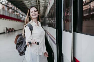 Happy beautiful young female passenger standing with luggage by train at railroad station - EBBF04147