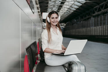 Zufriedene weibliche Passagierin hört Musik über Kopfhörer, während sie mit einem Laptop am Bahnhof sitzt - EBBF04150