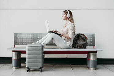 Junge Frau, die einen Laptop benutzt und über Kopfhörer Musik hört, sitzt auf einer Bank am Bahnhof - EBBF04152