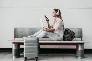 Junge Frau benutzt ihr Smartphone, während sie am Bahnhof über Kopfhörer Musik hört - EBBF04154