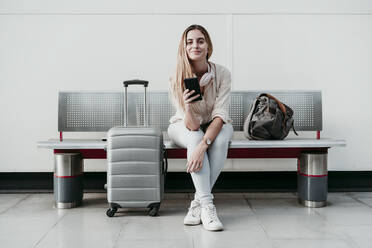 Beautiful woman holding smart phone while sitting with wheeled luggage at station - EBBF04155