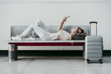 Young woman using smart phone while lying on bench at railroad station - EBBF04159