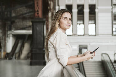 Blonde Frau schaut weg, während sie sich mit ihrem Smartphone am Geländer des Bahnhofs abstützt - EBBF04167